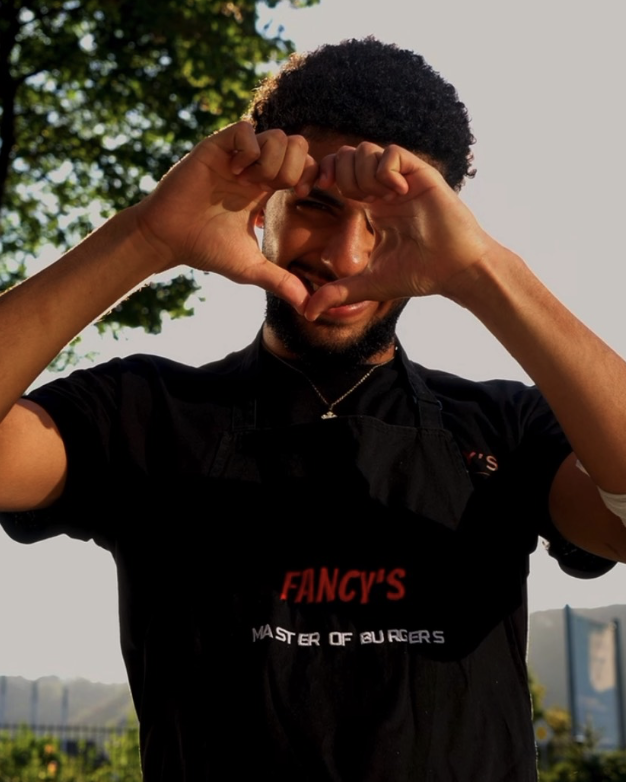 Minas formt mit beiden Händen ein Herz vor seinem Gesicht, im Fancy's Burger T-Shirt und schwarzer Schürze